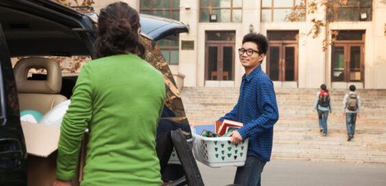 A young man smiling at his mom stands behind a minivan with an open trunk door, holding a laundry basket full of books.