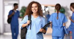 A smiling young woman wearing blue scrubs is holding a black clipboard and the strap of a backpack on her back.