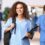 A smiling young woman wearing blue scrubs is holding a black clipboard and the strap of a backpack on her back.