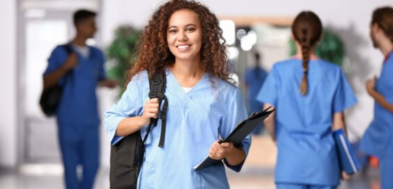 A smiling young woman wearing blue scrubs is holding a black clipboard and the strap of a backpack on her back.
