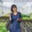 Smiling person in a blue apron uses a tablet among raised beds of green and red leafy plants inside a greenhouse.