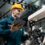 A man in a yellow hard hat and a blue one-piece work suit examines the robotic arm of a machine while holding a tablet.