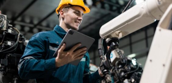 A man in a yellow hard hat and a blue one-piece work suit examines the robotic arm of a machine while holding a tablet.