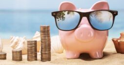 A pink piggy bank wearing sunglasses sits on a sandy surface next to growing piles of coins. Shells are next to the pig.