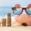 A pink piggy bank wearing sunglasses sits on a sandy surface next to growing piles of coins. Shells are next to the pig.