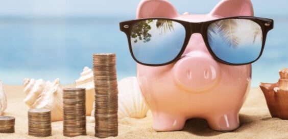 A pink piggy bank wearing sunglasses sits on a sandy surface next to growing piles of coins. Shells are next to the pig.