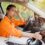A person wearing an orange shirt dabbing their head with a napkin while sitting in the driver’s seat of car.