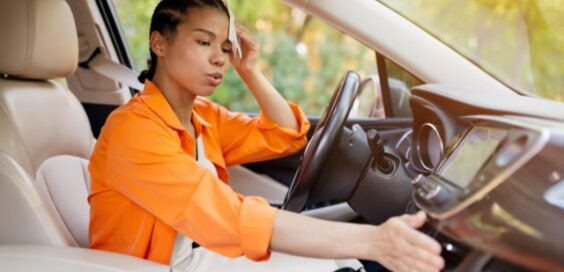 A person wearing an orange shirt dabbing their head with a napkin while sitting in the driver’s seat of car.
