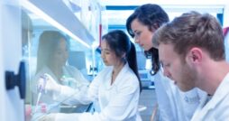 Three people in white lab coats gather around a workbench as one uses a pipette behind glass in a laboratory.