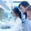 Three people in white lab coats gather around a workbench as one uses a pipette behind glass in a laboratory.