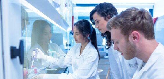 Three people in white lab coats gather around a workbench as one uses a pipette behind glass in a laboratory.