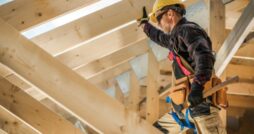 A worker wearing a yellow hard hat and tool belt standing among wooden beams while holding a hammer.