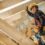 A worker wearing a yellow hard hat and tool belt standing among wooden beams while holding a hammer.