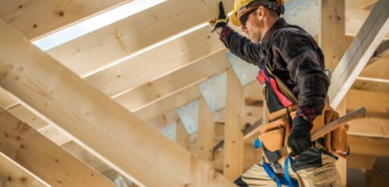 A worker wearing a yellow hard hat and tool belt standing among wooden beams while holding a hammer.