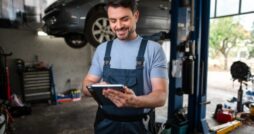A smiling man in blue coveralls using a tablet near a raised car and tools inside an auto repair shop.