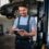 A smiling man in blue coveralls using a tablet near a raised car and tools inside an auto repair shop.