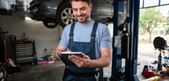 A smiling man in blue coveralls using a tablet near a raised car and tools inside an auto repair shop.