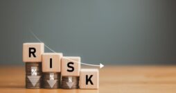 A close-up of four wooden blocks with letters on descending stacks of coins. The letters spell out "RISK."