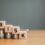 A close-up of four wooden blocks with letters on descending stacks of coins. The letters spell out "RISK."