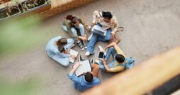 Downward view of five students sitting in a circle on the floor. They have notebooks, a laptop, and a tablet.