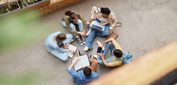 Downward view of five students sitting in a circle on the floor. They have notebooks, a laptop, and a tablet.