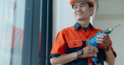 A man wearing an orange hard hat and work uniform holding a blue power drill while standing beside a large window.