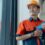 A man wearing an orange hard hat and work uniform holding a blue power drill while standing beside a large window.