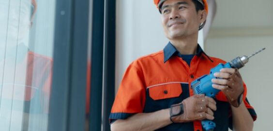 A man wearing an orange hard hat and work uniform holding a blue power drill while standing beside a large window.