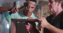 A young man using a hammer on a wooden project while an older man guides him in a carpentry workshop setting.