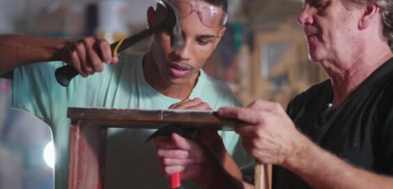 A young man using a hammer on a wooden project while an older man guides him in a carpentry workshop setting.