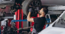 A man wearing gloves and dark work clothes holding a car wheel beneath a raised silver vehicle inside an auto garage.