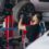 A man wearing gloves and dark work clothes holding a car wheel beneath a raised silver vehicle inside an auto garage.
