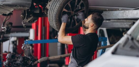 A man wearing gloves and dark work clothes holding a car wheel beneath a raised silver vehicle inside an auto garage.