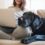 A woman sits cross-legged on a couch while holding a laptop. A black dog lies beside her with its head on her knee.