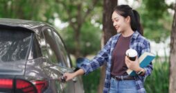 A woman in a plaid shirt holds a takeaway coffee cup and a blue book while she opens the door of a gray car outdoors.