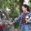 A woman in a plaid shirt holds a takeaway coffee cup and a blue book while she opens the door of a gray car outdoors.