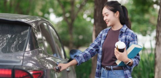 A woman in a plaid shirt holds a takeaway coffee cup and a blue book while she opens the door of a gray car outdoors.