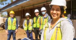 A close-up of a woman in a hard hat standing in front of people wearing safety gear in an outdoor area.