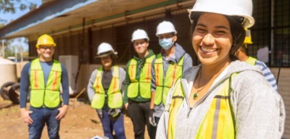 A close-up of a woman in a hard hat standing in front of people wearing safety gear in an outdoor area.