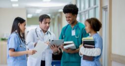 A doctor in a white coat shows a tablet to three medical colleagues wearing scrubs and holding documents in a hospital.
