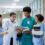 A doctor in a white coat shows a tablet to three medical colleagues wearing scrubs and holding documents in a hospital.