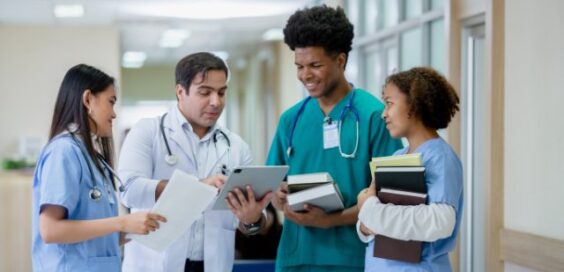 A doctor in a white coat shows a tablet to three medical colleagues wearing scrubs and holding documents in a hospital.