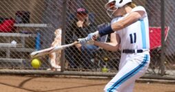 A young female softball player in a white and blue uniform and a white batting helmet is swinging at a softball pitch.