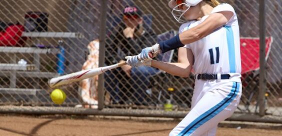 A young female softball player in a white and blue uniform and a white batting helmet is swinging at a softball pitch.