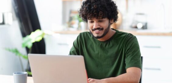 A young man wearing a green T-shirt sits at a table typing on a silver laptop with an open book and a white mug next to him.